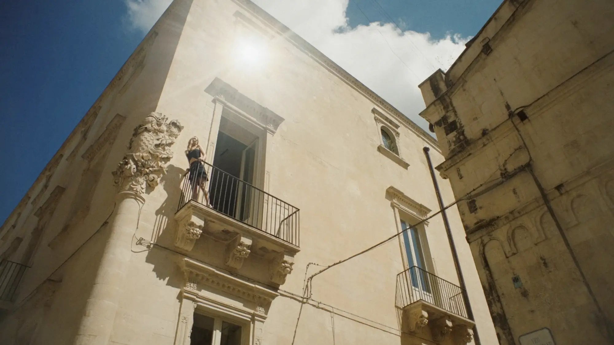 Sunlit Mediterranean heritage building with an ornate stone balcony and a woman standing by the railing under a bright blue sky.