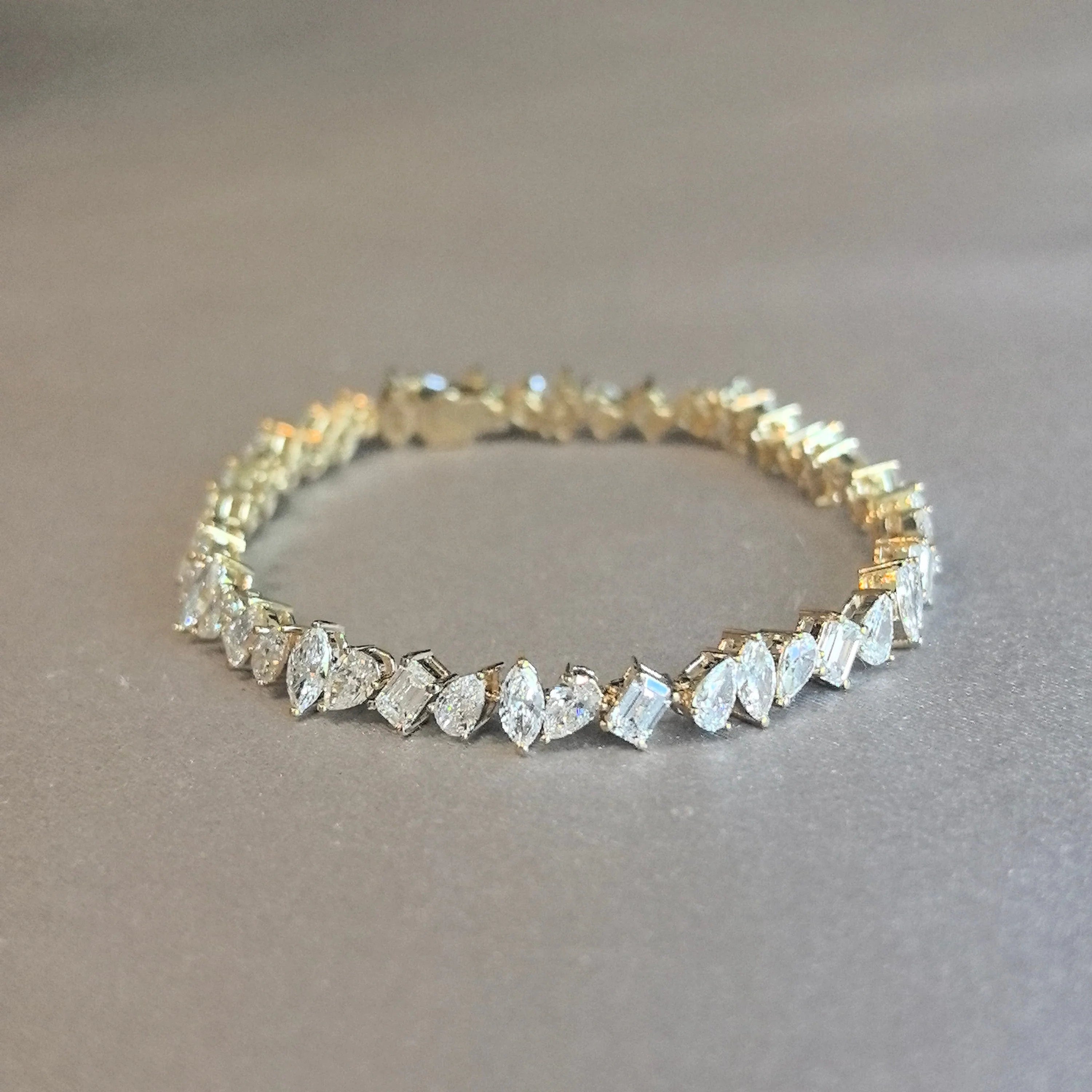 Gold tennis bracelet featuring mixed-cut diamonds in marquise, pear, emerald, and round shapes, displayed on a soft gray surface.