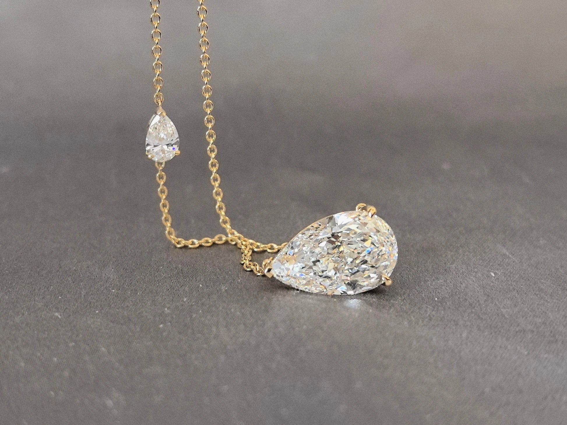 Gold lariat necklace featuring two pear-shaped diamond pendants on a delicate chain, displayed on a gray fabric surface.